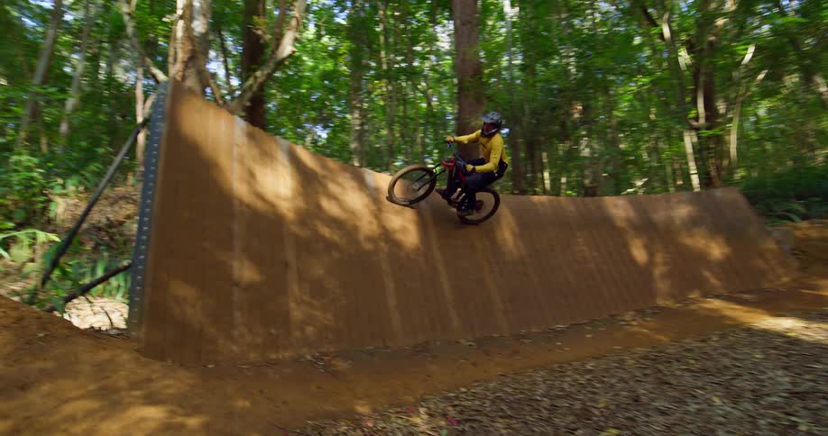 A mountain biker rides skillfully along a dirt trail in a sunny forest executing a jump off a large wooden ramp before continuing down the path