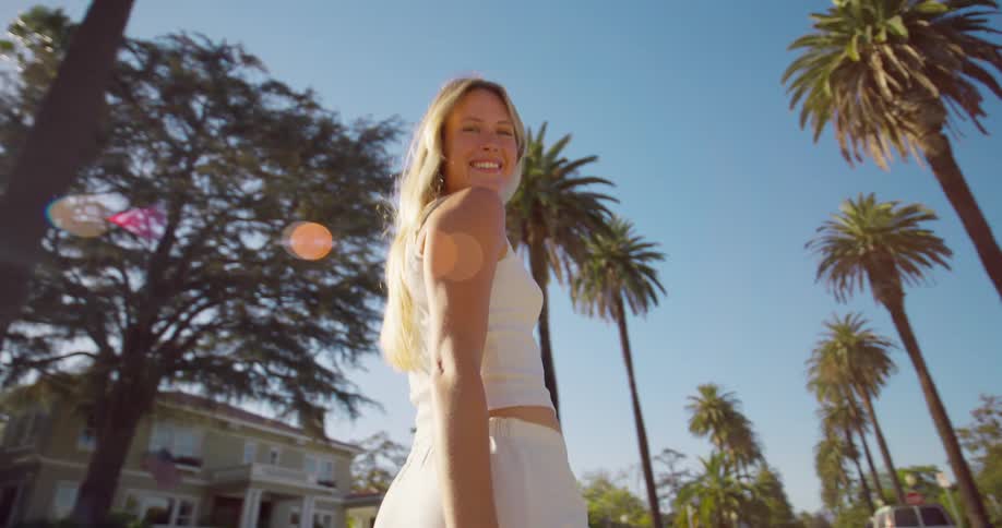 A happy woman with long blonde hair walks down a sunny street lined with palm trees turning back to smile at the camera