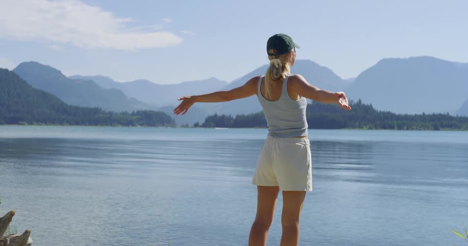 Woman with open arms gazes at a lake surrounded by mountains