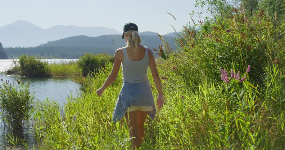 A woman wearing a cap walks through tall grass at the edge of a large lake