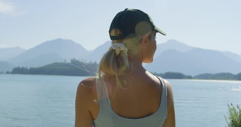 The video shows a woman from behind looking out at a large body of water in the background there are mountains under a clear sky