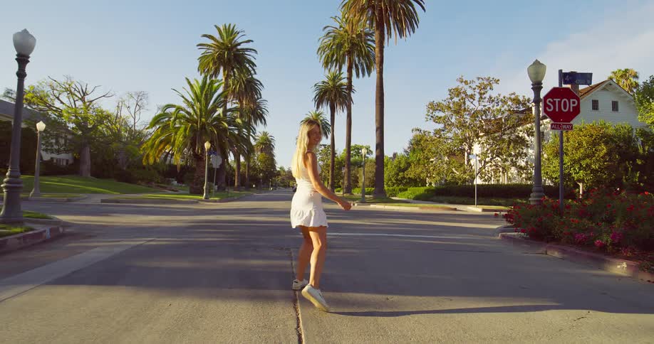 A video shows a young woman with long blonde hair dressed in a white dress walking down a street lined with palm trees she turns and smiles at the camera then twirls and dances happily