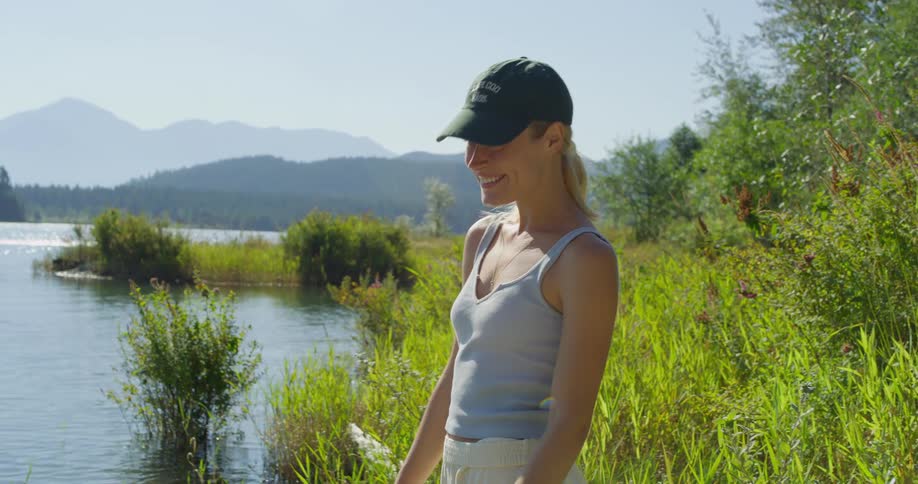 A medium shot shows a young woman standing by a lake on a clear day smiling she is wearing a green baseball cap and a light blue tank top with mountains visible in the distance