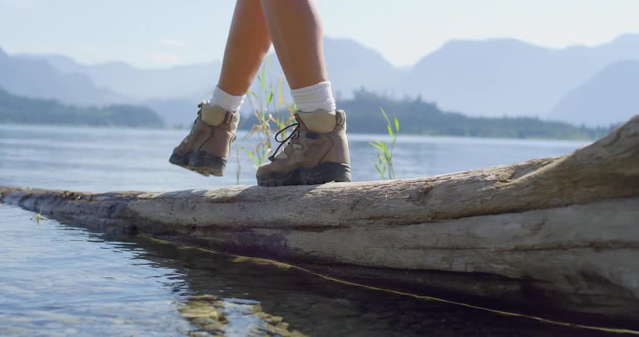 Person walking on a log by a lake wearing hiking boots and white socks with mountains in the background