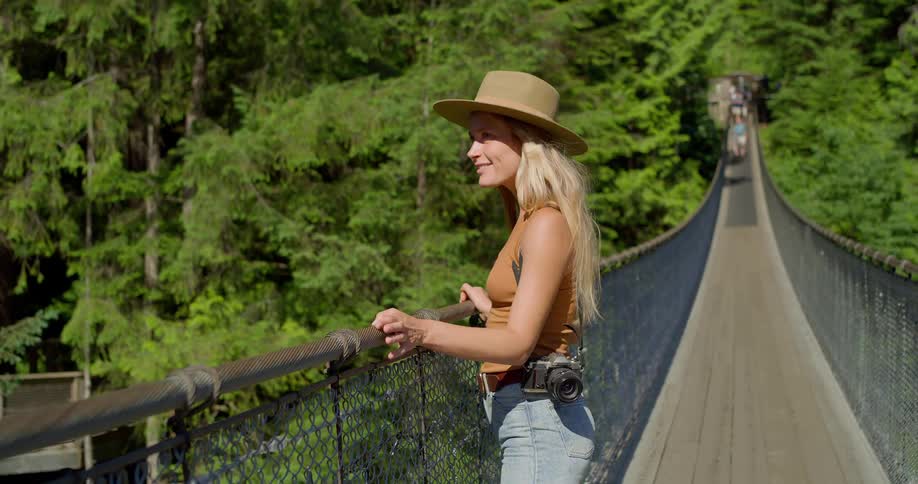 Woman wearing a hat with a camera on her waist looks at the scenery on a suspension bridge surrounded by forest