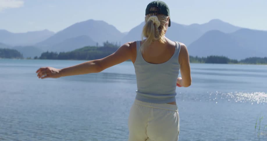 A woman with a ponytail and a baseball cap is jogging joyfully near a scenic lake with mountains in the background smiling as she moves