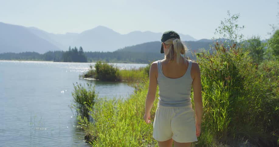A woman lingers by a bright lake shoreline mountains hazy beyond and tall grasses lit by summer sun