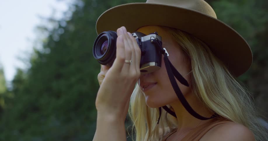 A young blonde woman is outside wearing a hat and holding a camera up to her face to take a picture after a moment she lowers the camera and looks up thoughtfully