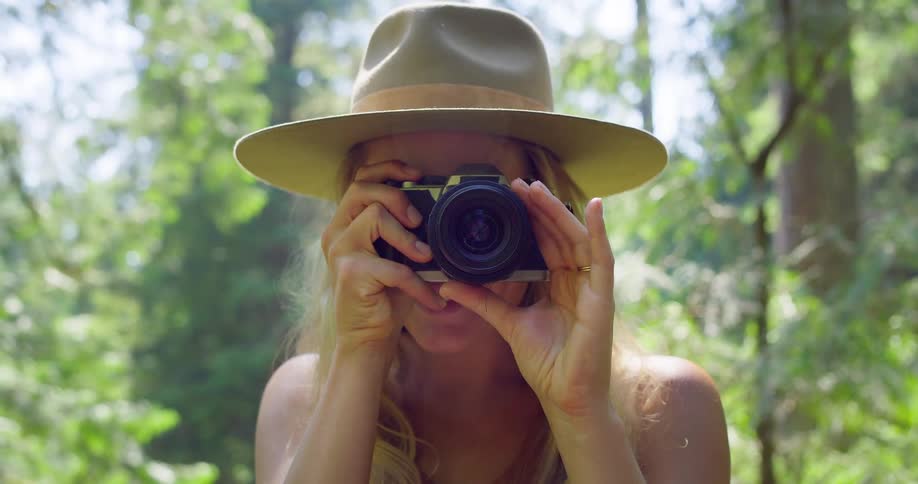 The video shows a woman wearing a hat standing outdoors in a forest she looks at the camera and then lifts a camera to her face to take a picture