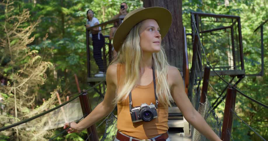 A woman wearing a hat and camera explores a forest canopy bridge