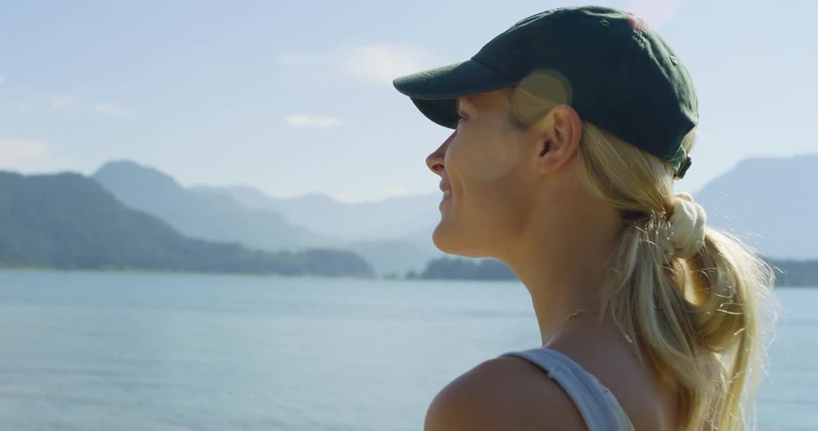 A young woman standing on the shore watching the lake wearing a hat and smiling at midday