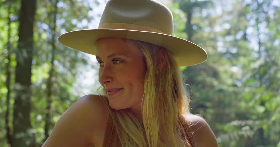 A happy woman wearing a tan hat is looking up and smiling in a sunlit forest