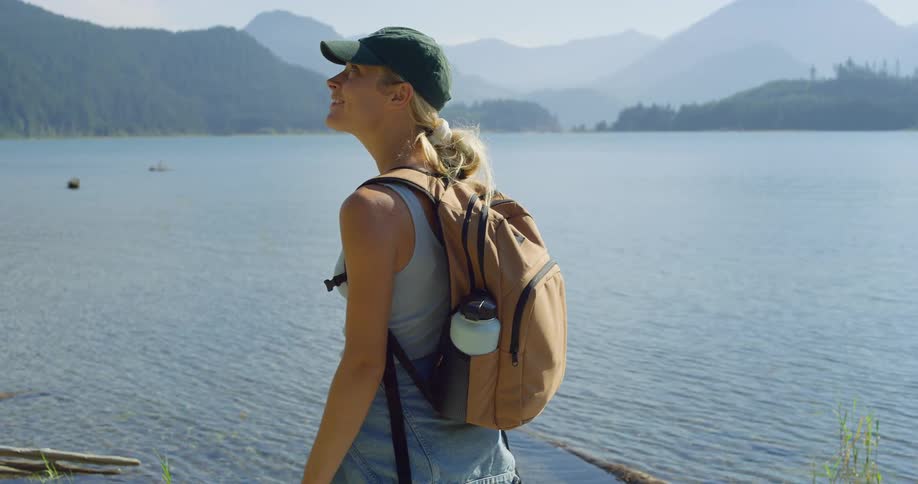 A blonde woman wearing a cap and with a beige backpack on her back is already looking at a huge lake in front of her