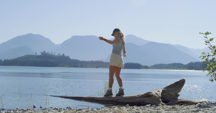 A woman in hiking boots carefully balances and walks along a large piece of driftwood on a lake shore with a scenic mountain range in the background