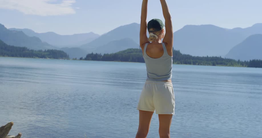 Happy woman stands by a lake stretches her arms up then turns and walks away