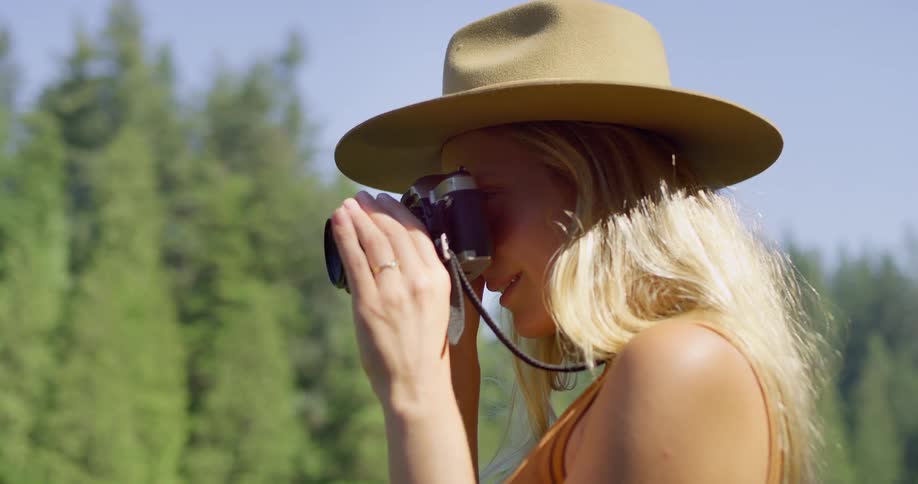 Sideprofile closeup of a woman in a tan hat lifting a small camera to her eye with soft sunlight and trees out of focus behind her