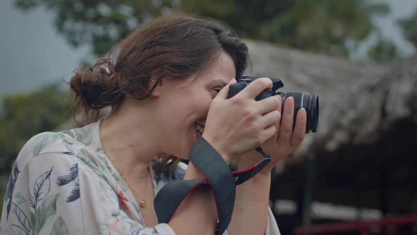 A woman with brown hair tied up in a bun is holding a camera with a black lens strap she is wearing a floral shirt and a gold necklace she smiles slightly as she looks through the viewfinder appearing focused on taking a picture