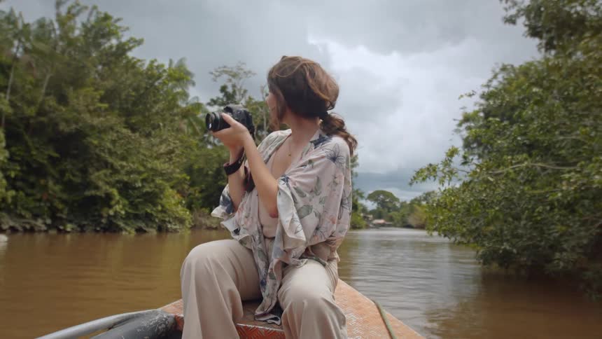 The video shows a woman sitting in a boat on a murky river surrounded by trees she is holding a camera up to her face appearing to take a photo of something out of frame