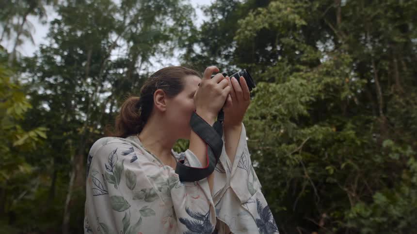 A woman in a floral shirt standing in a forest holding a professional camera and looking through the viewfinder to take a picture
