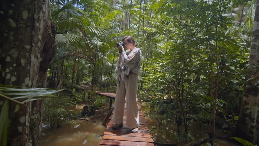 Female photographer stands on a walkway above the water in the woods while taking a picture