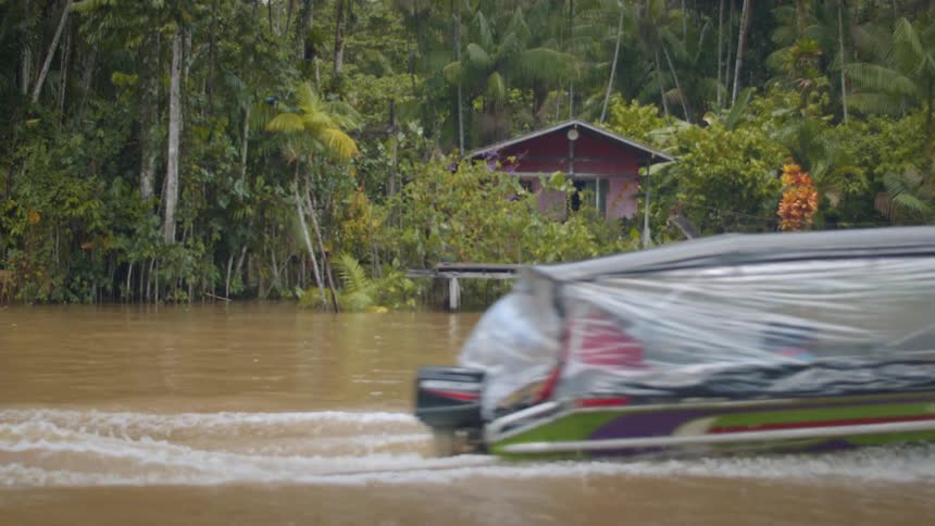 A colorful longboat speeds past a stilt house along a muddy tropical river
