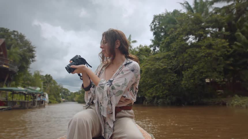 A young female photographer takes photos with a camera while siting on a canoe in a river during daytime