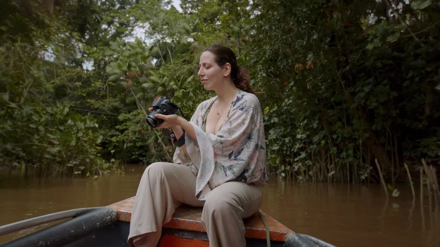 The video shows a woman sitting in a small boat on a murky river surrounded by dense jungle foliage she is holding a camera taking pictures of the scenery and looking around at her surroundings