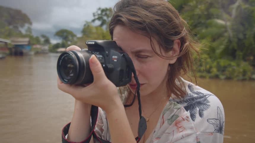 A woman with tousled brown hair wearing a floral patterned shirt holds a camera up to her eye seemingly about to take a photo on a river