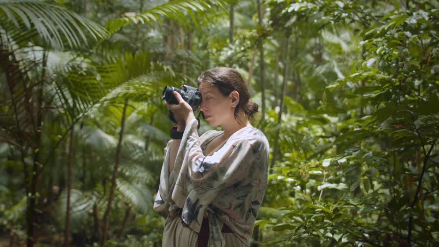 A woman is in a lush jungle taking photographs with a camera
