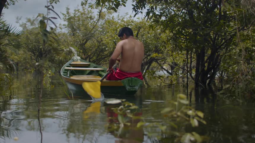 Shirtless man wearing red shorts paddles a green and yellow canoe on calm waters surrounded by trees