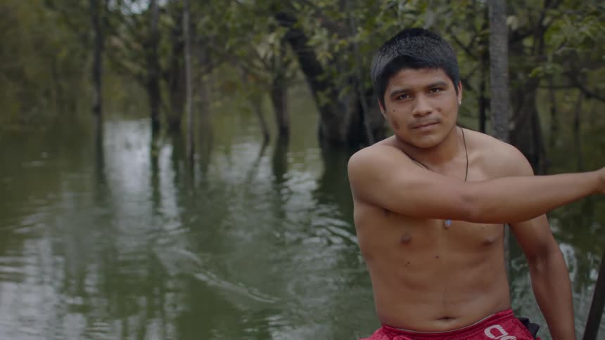 A young shirtless man paddles a boat with a single oar navigating through the calm waters of a flooded forest or jungle river while looking at the camera