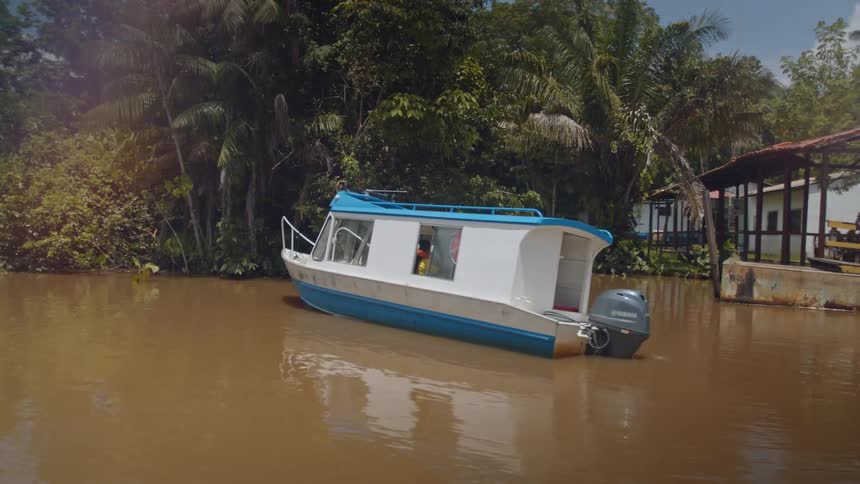 White and blue boat docked on a dark river next to dense vegetation and simple wooden structures