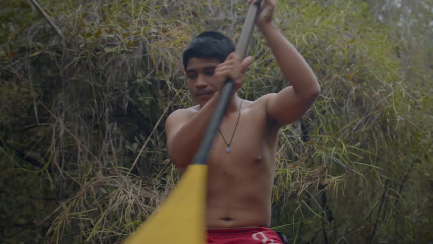 A man with a shaved head is seated in a canoe using an oar to row through a river surrounded by lush green trees and brush