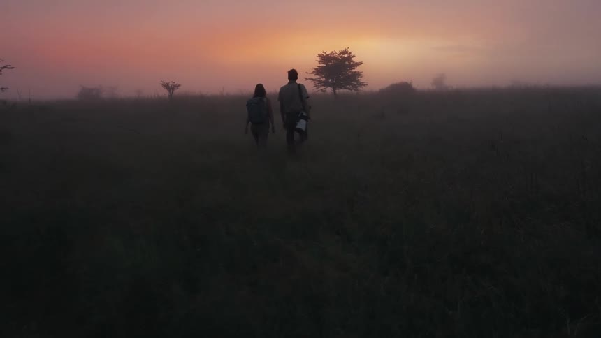 Two people with backpacks walk side by side across a foggy field at sunrise with a single tree on the horizon and the sky glowing with shades of pink and orange