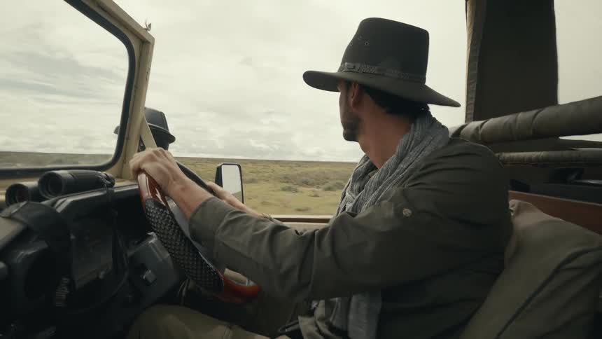 The face of a man wearing a hat driving an opentopped car in a savannah