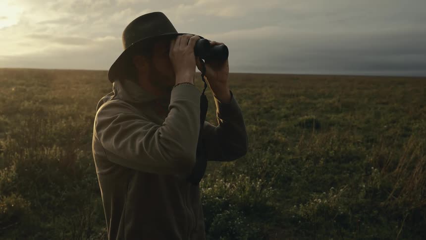 A man uses binoculars to scan the horizon in a vast grassy field at sunset