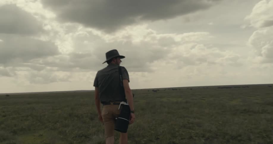 A man stands with his back turned walking in a large field of grass on a day with heavy clouds in the sky