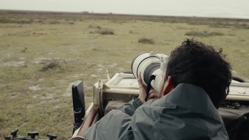 A person with a large telephoto lens in the back of a safari vehicle looking out across a vast grassy plain