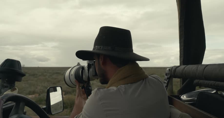 A man wearing a hat is sitting in a vehicle and holding a large camera with a telephoto lens he is looking through the viewfinder possibly photographing something in the distance