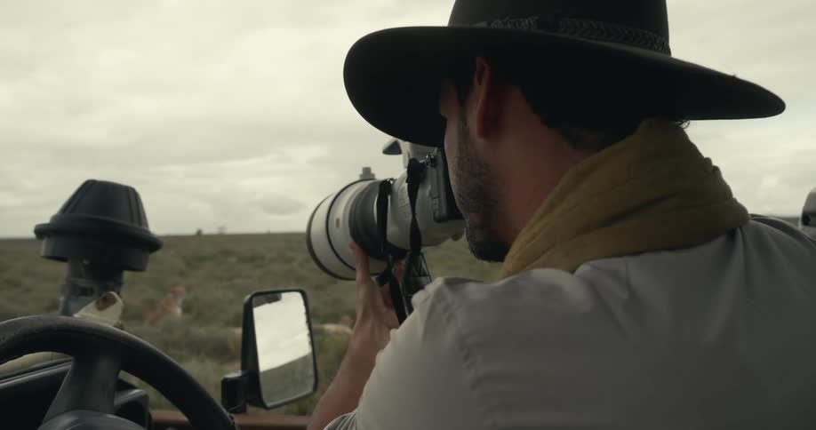 A man sitting in an openroof truck using a digital camera to photograph the wildlife at midday wearing a hat and a scarf around his neck