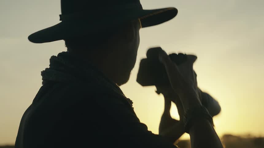 A closeup backlit shot of a person in a hat and scarf is shown with the sun setting behind them the person brings a professional camera with a telephoto lens to their eye to take a photo