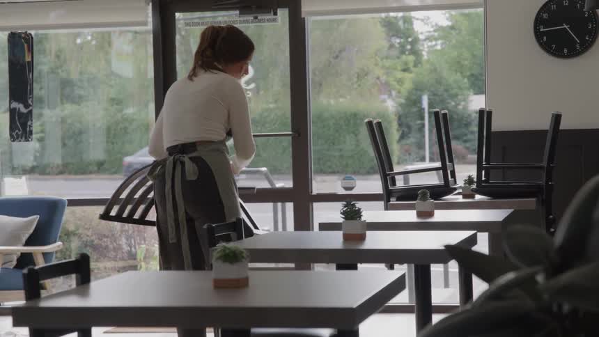 The video shows a worker setting up a cafe for business turning over chairs that were stacked on top of the tables the cafe appears to be empty with a large clock on the back wall