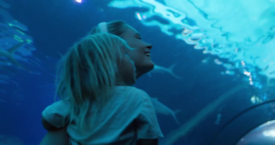 A blonde mother holds her young child pointing up in the blue lit aquarium as fish swim past them