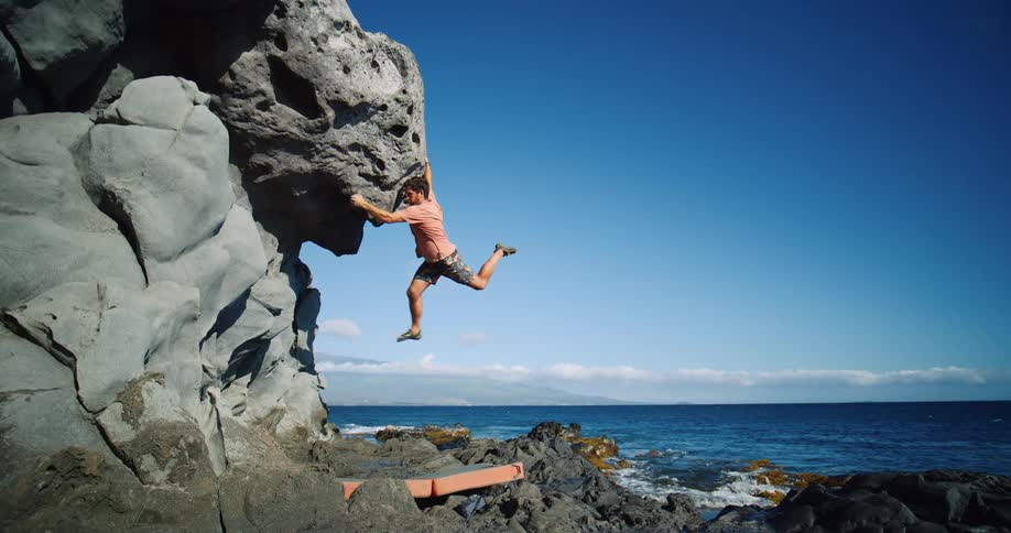 A climber hangs on a bulging coastal boulder with waves crashing below