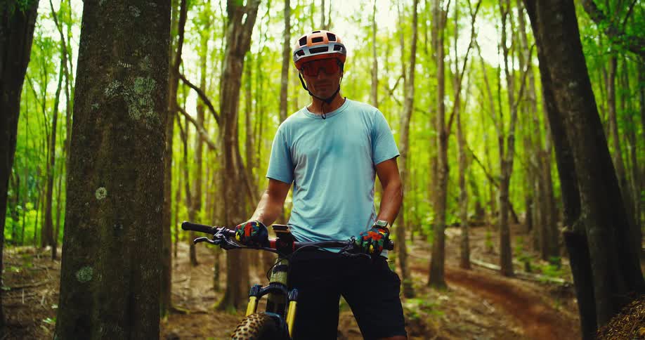 The video shows a male mountain biker in a forest holding onto his bike he is wearing a helmet and gloves and is looking at the camera