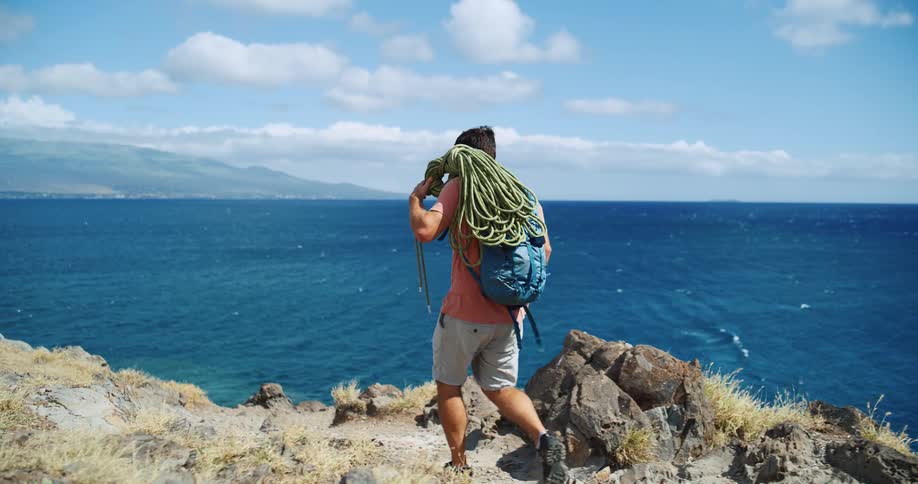 The video shows a man standing on a rocky cliff overlooking the ocean he is wearing a backpack and has a large bundle of climbing rope slung over his shoulder