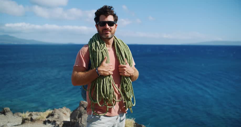 A man poses on a cliff over the ocean with climbing ropes over his shoulders