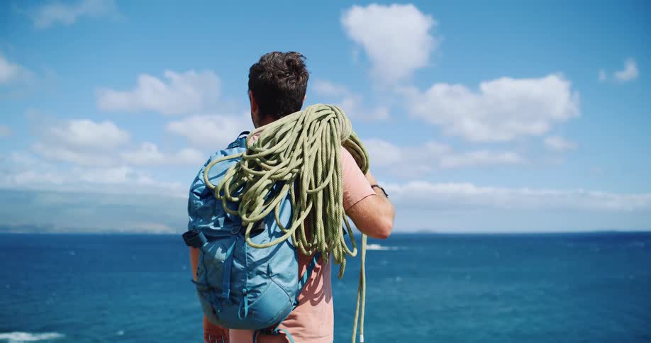 A man with his back turned is standing with a blue backpack and holding a rope on his shoulder looking at a blue ocean