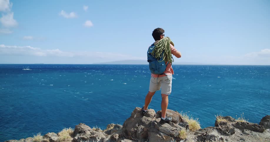 Man with a backpack and climbing rope looks at the sea from the top of a cliff