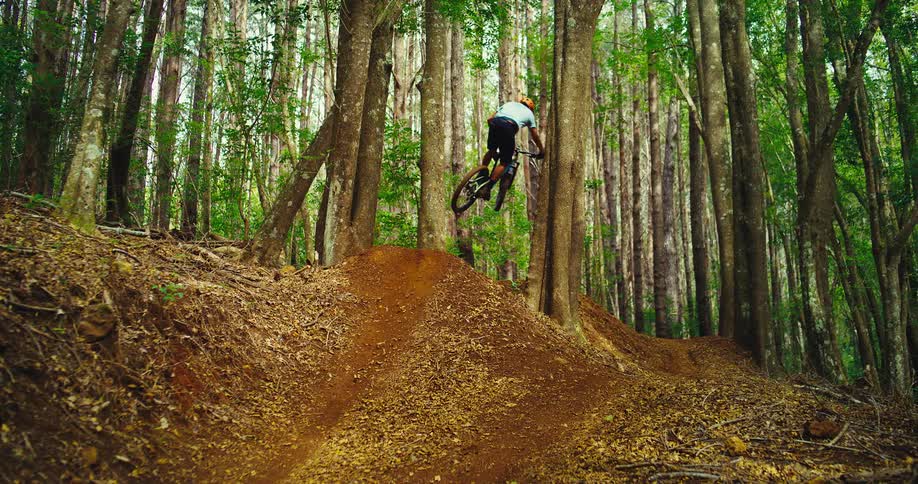 The video shows a mountain biker riding on a dirt trail through a forest the biker goes over a small jump and the camera follows from behind as he continues down the trail